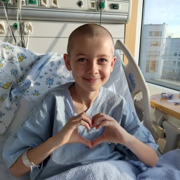 A young cancer patient smiling and making a heart shape with their hands in a hospital bed
